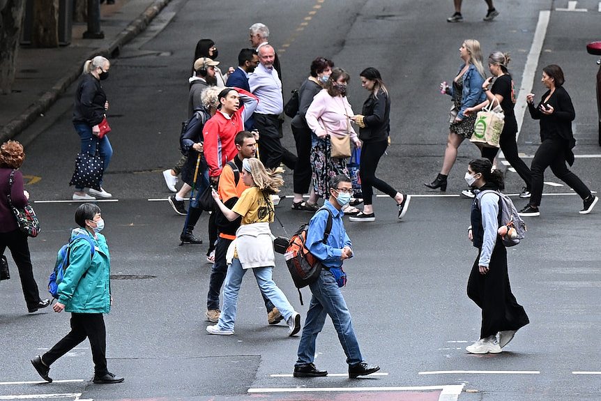 Many people, some wearing face masks, walk across a road.