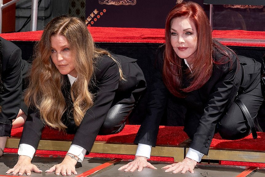 Two women with long red hair are immersing their hands in wet cement on a footpath.