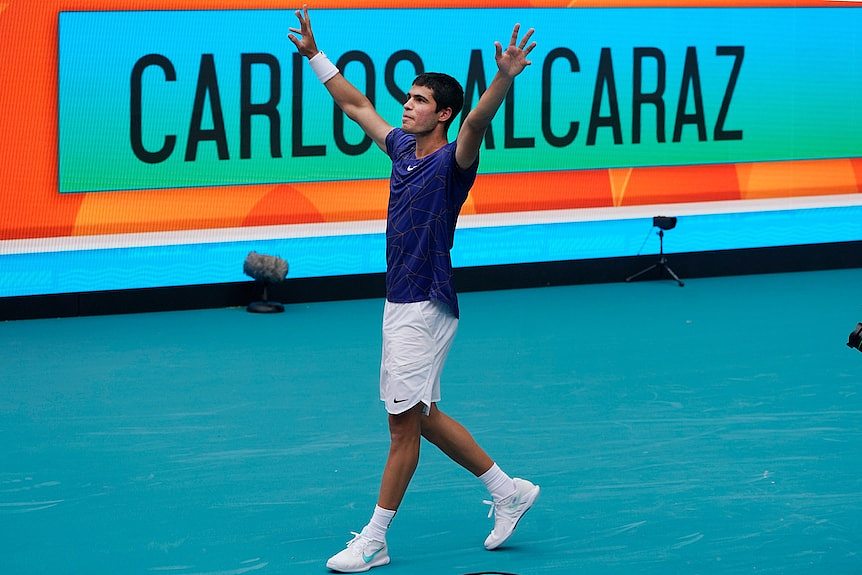 Spanish tennis star Carlos Alcaraz raises his arms in triumph as he walks with a 'Carlos Alcaraz' sign behind him. 