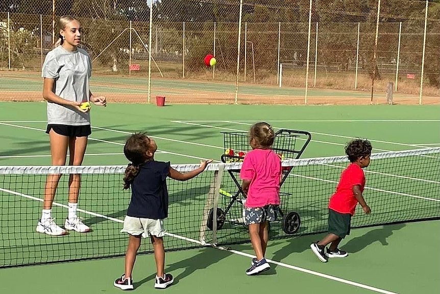 A young woman coaches three small children on a green tennis court