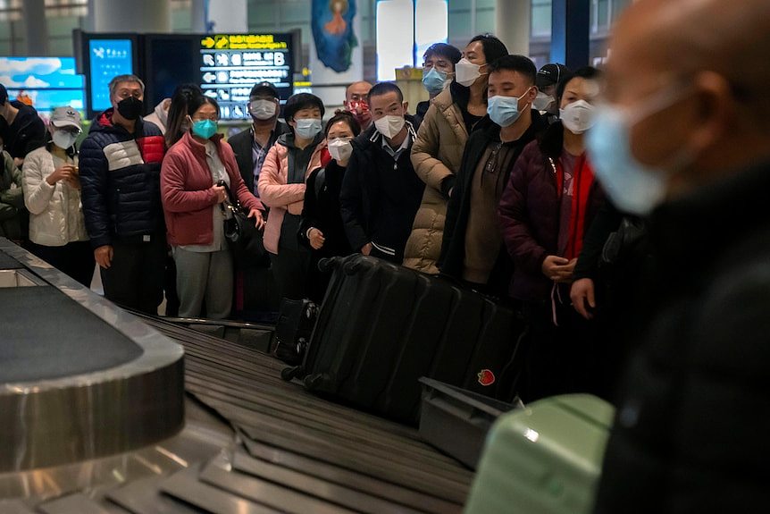 Travellers wearing face masks wait for their luggage to come along a conveyor belt.