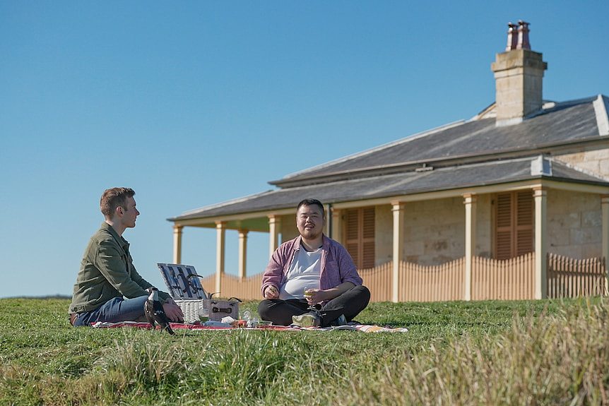 Two men sit on a grassy hill for a picnic. A cottage house is behind them.