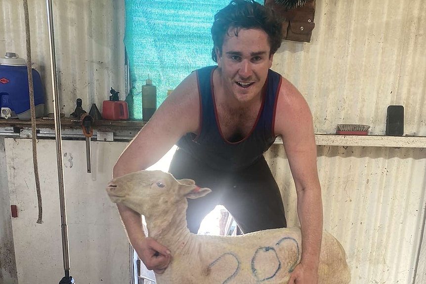 A young man in a singlet smiles while he holds a sheep with the number 200 painted across its body.