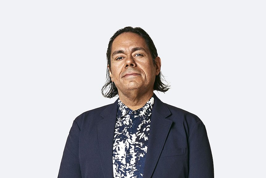 William Barton is an Indigenous man with brown skin and mid-length dark hair. He is wearing a dark blue jacket.