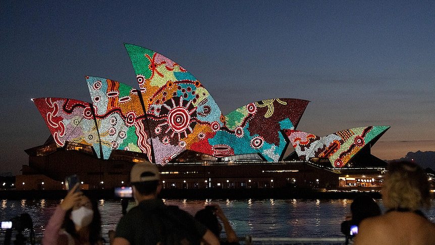 The Sydney Opera House is lit up with Indigenous artwork. It is twilight and onlookers can be seen taking in the view