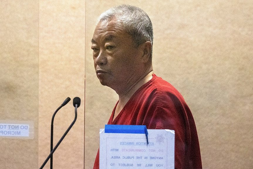 A man in a red jumpsuit appears behind a glass screen. 