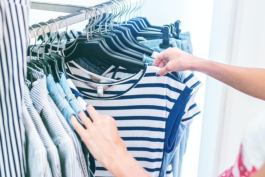 a shopper browsing through a rack of clothes stops to look closely at a t-shirt