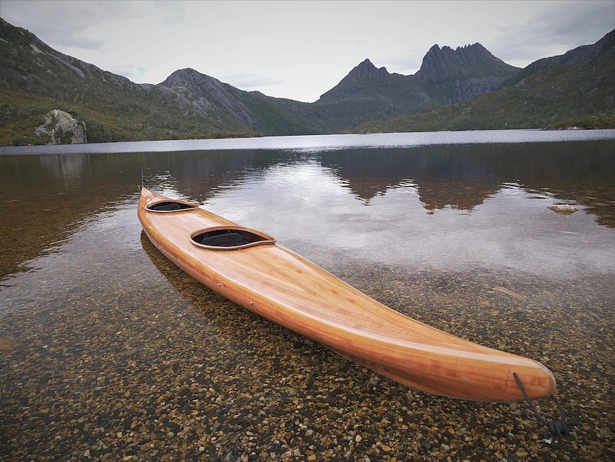 A timber kayak in the shallows of a lake, mountains looming behind.