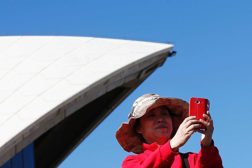 一个穿亮红色衣服的亚洲女人在悉尼歌剧院前自拍。