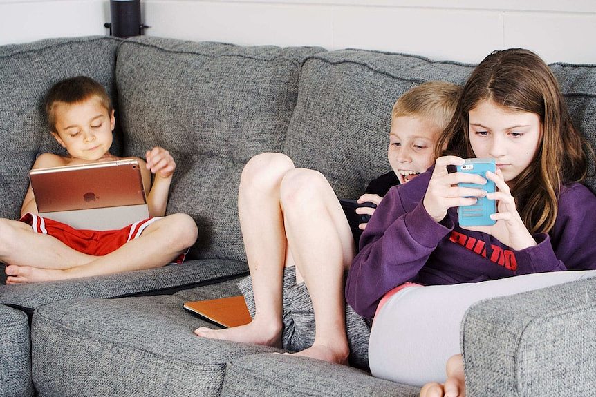 Three young children sit on a couch engrossed in phones and tablets.