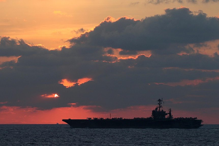 A large ship crosses a flat ocean with the sunsetting behind clouds in the background.