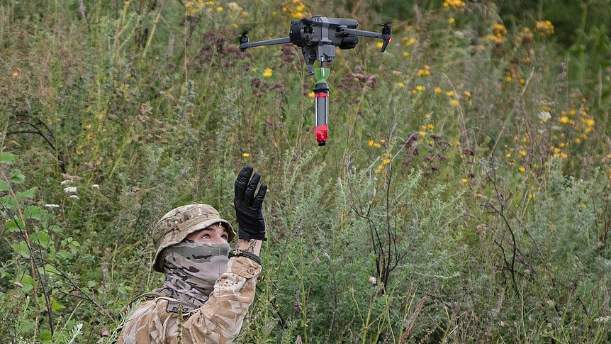 A soldier raises their hand near a small commercial drone holding an explosive. 