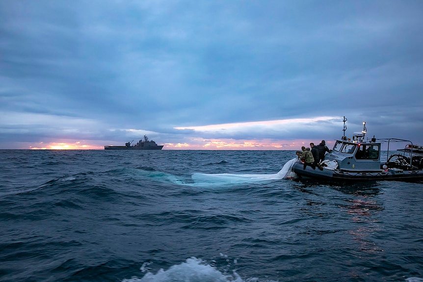 Three boats in the ocean, one with crew pulling a plastic sheet with sunrise in the background