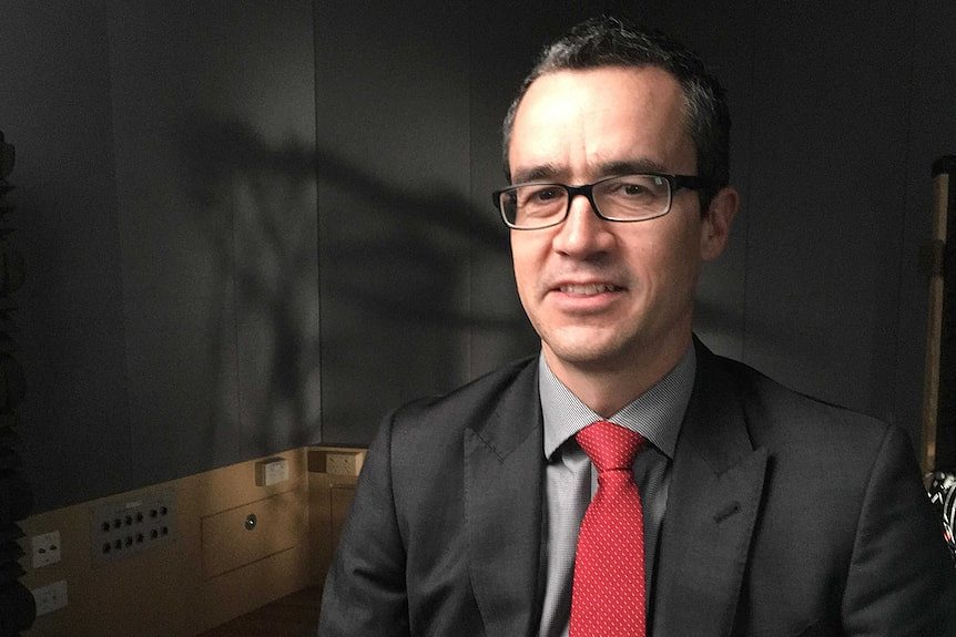 A slightly smiling middle-aged man wears black suit, grey shirt, red tie. Glasses, dark background, shadows on the wall.