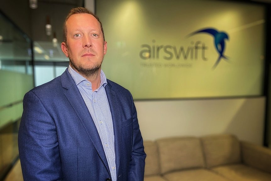 A serious middle-aged man in blue suit, white and blue patterned shirt in front of a wall with airsiwft and its logo, a couch.