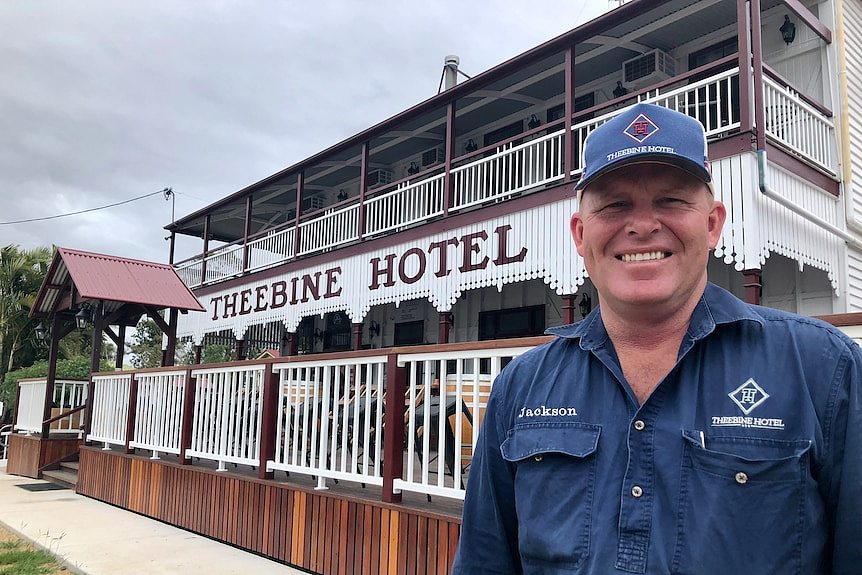 A man in a cap smiles at the camera with the Theebine hotel and new front deck behind him.