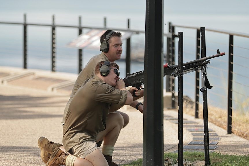 Two soldiers kneeling behind a mock machine gun and pointing it at the sky, on pavement next to a rail overlooking the ocean.