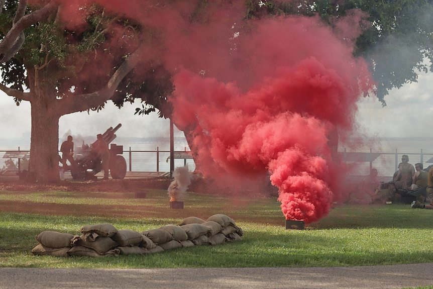 A cloud of red smoke billows from a small container on the grass on the Darwin Esplanade as soldiers watch on.