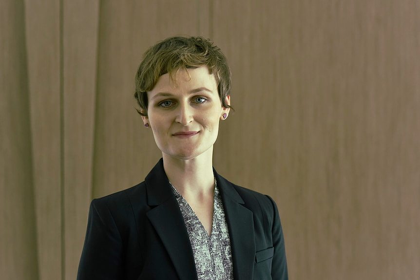 A white woman with shortish wavy hair stands, wearing a business jacket