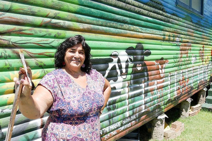 Woman stands in front of her mural of cows in green paddock.