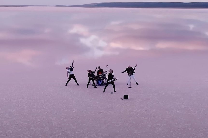 Wide aerial shot of a band standing on dried, pink salt lake with cloudy pin skies reflected  