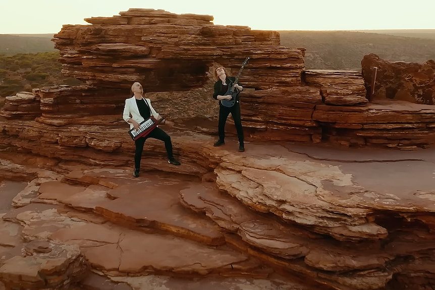 Man in white jacket plays keytar, man in black shirt plays guitar with window-like rock formation in background