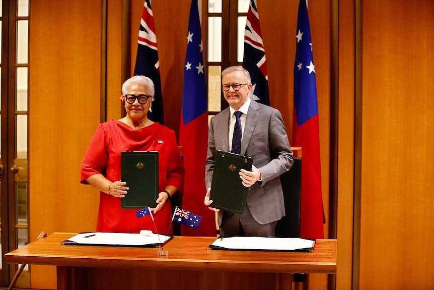 A woman and a man display leatherbound formal documents as they stand before Samoan and Australian flags.