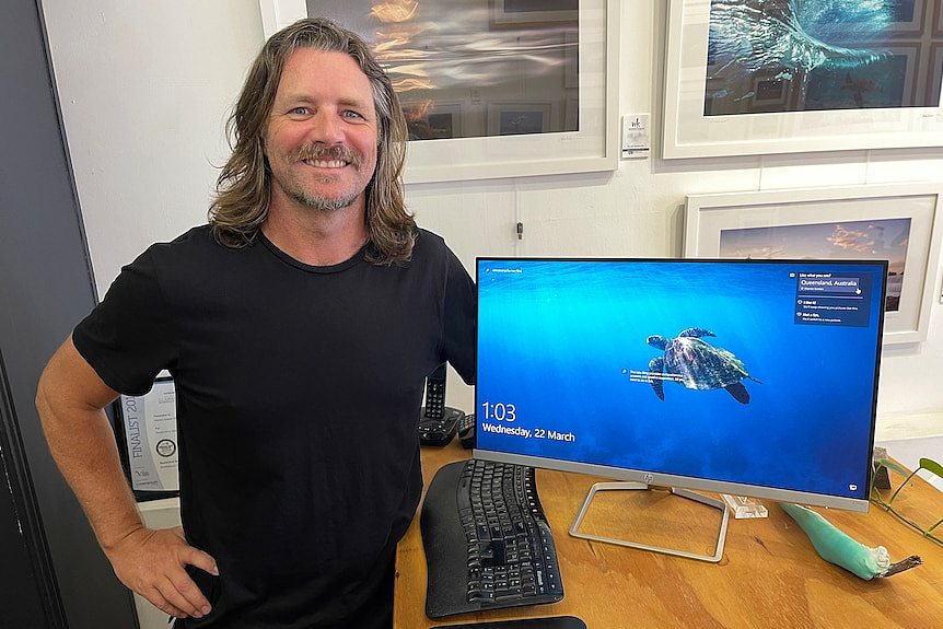 Warren Keelan stands in a black t-shirt in front of a computer monitor with a picture of a turtle on it.