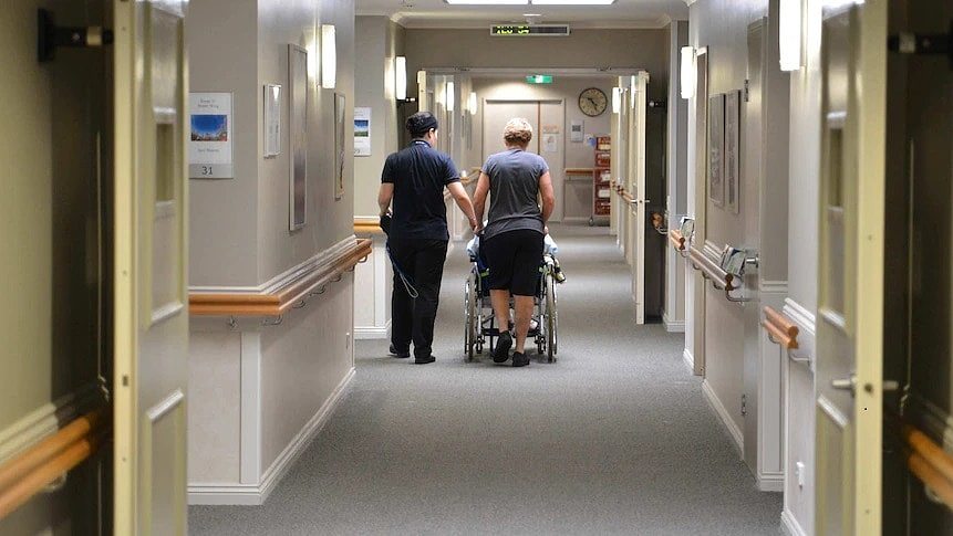 Aged care workers accompany a resident down the hallway of their facility