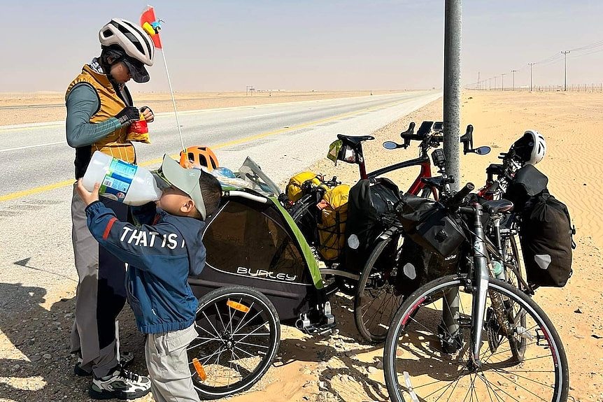 A woman and a young boy stop beside their bike next to a bitumen road through a sandy desert. 