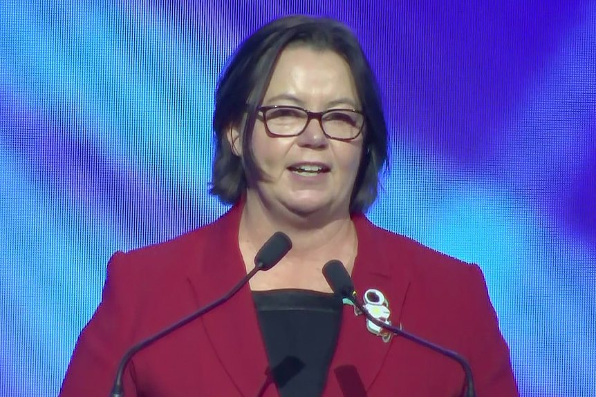 Madeleine King delivering a speech in a red jacket and black shirt at a podium on a stage in front of a blue electronic screen.