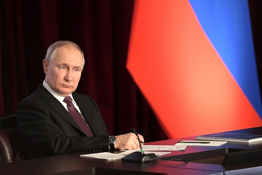 Russia's President Vladimir Putin sits at a desk. He has a blank look on his face.