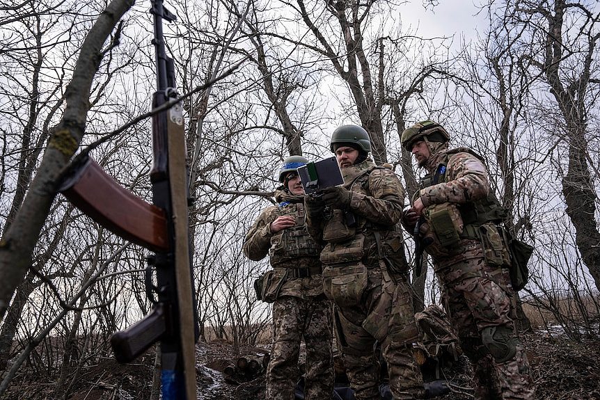 Soldiers gather in a stand of leafless trees to watch a drone operator in action with a smart device.