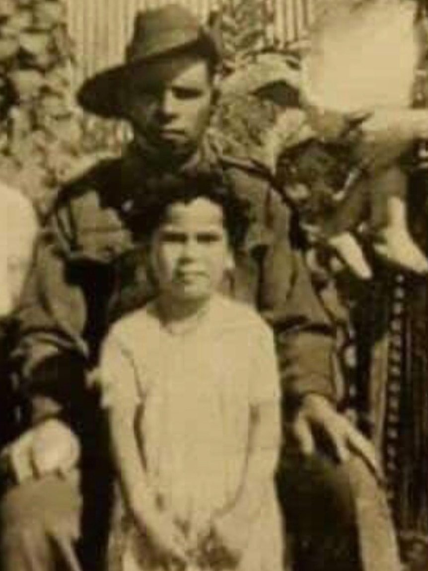 A sepia toned photo shows a young girl posing for a photo in front of a man wearing an army uniform and hat