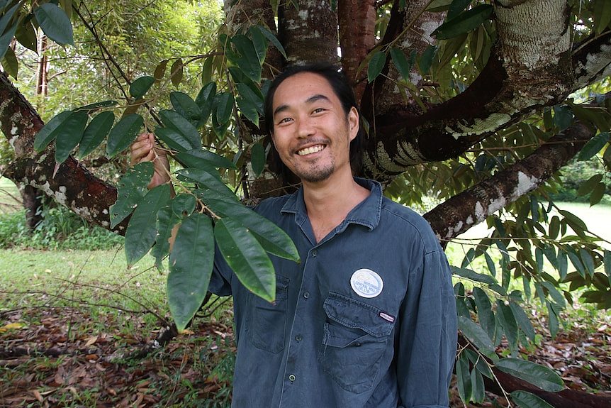A man in a blue work shirt stands under a durian tree, holding some leaves and smiling at the camera.