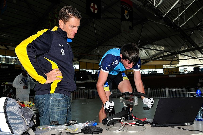 A man watches as a young cyclist rides on a stationary bike.