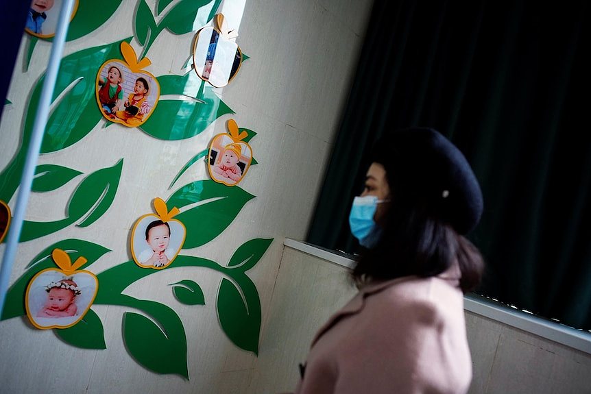 a woman in a face mask walks past a images of children on a wall inside a hospital