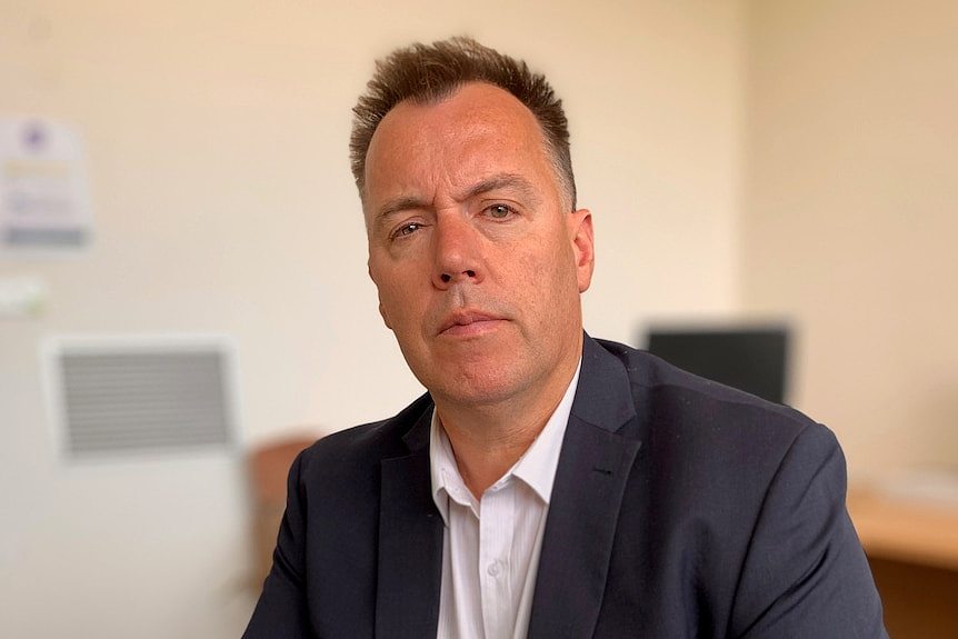 A man in a suit jacket sits in an office, with a serious expression.