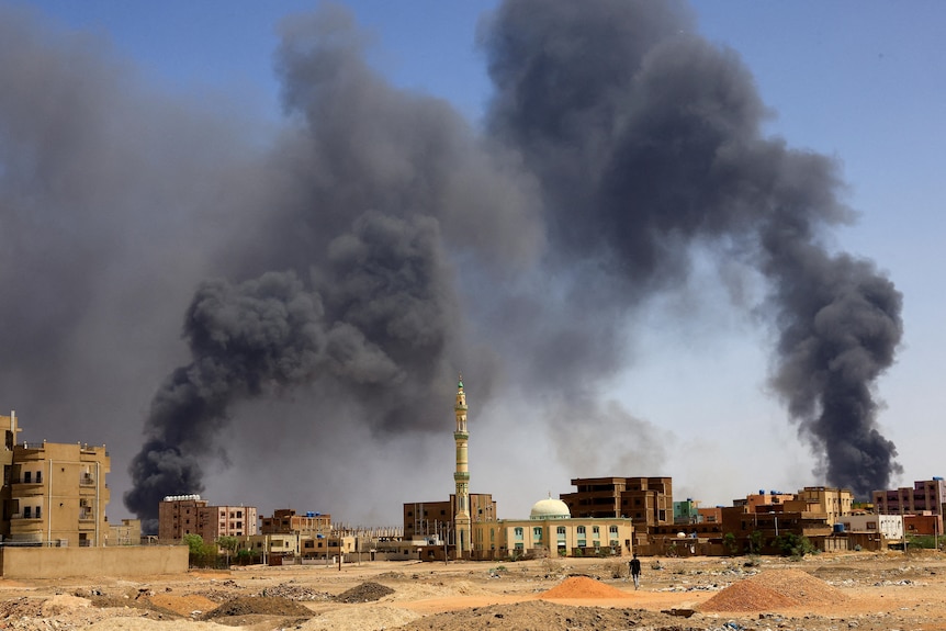 喀土穆市的建筑物中冒出浓烟，可以看到前景中的一名男子在地上行走。