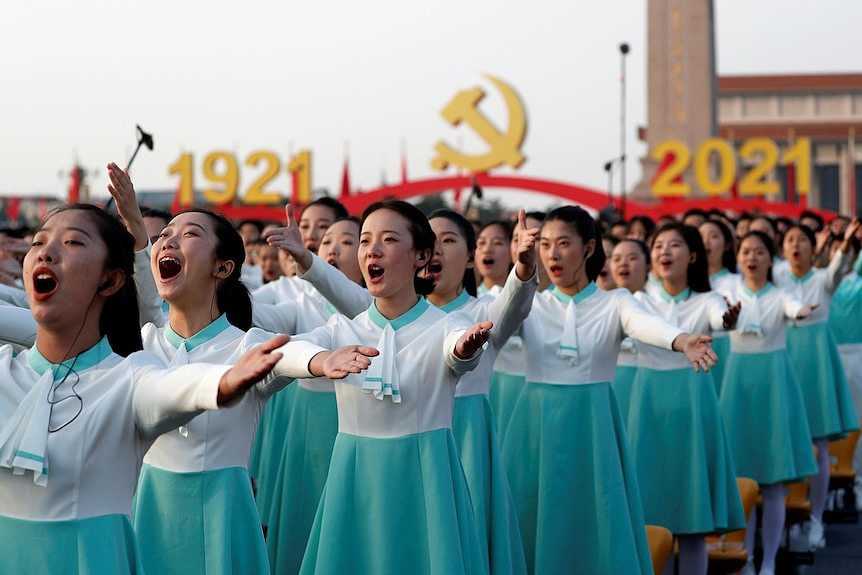 A group of Chinese women in blue skirts singing with their arms raised.