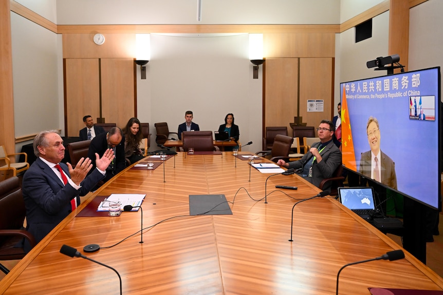 A suited man raises both hands as he speaks to another man via a TV video link, with others observing while sitting at a table.
