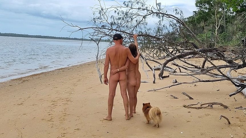 一个裸体男人在海滩上搂着一个裸体女人。 一只小狗站在他们身后。