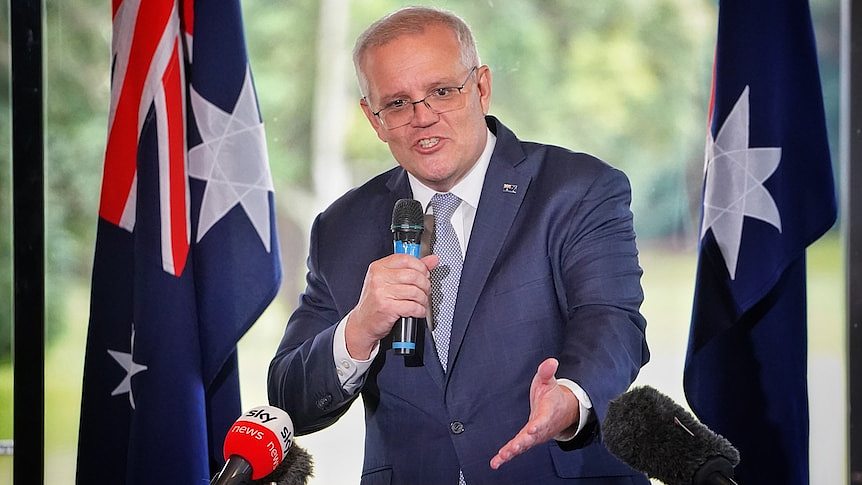 Morrison holds a microphone as he speaks from a lectern, with Australian flags beside him.