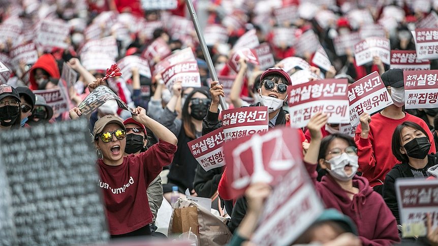 A large crowd of women wearing red hold posters and placards with Korean writing. Some are shouting.