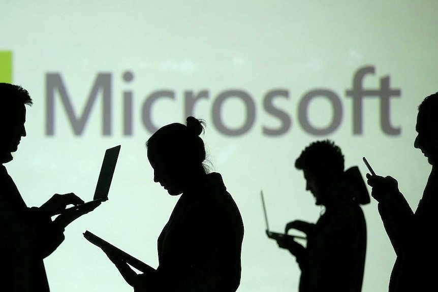 Silhouettes of laptop and mobile device users are seen next to a screen projection of Microsoft logo