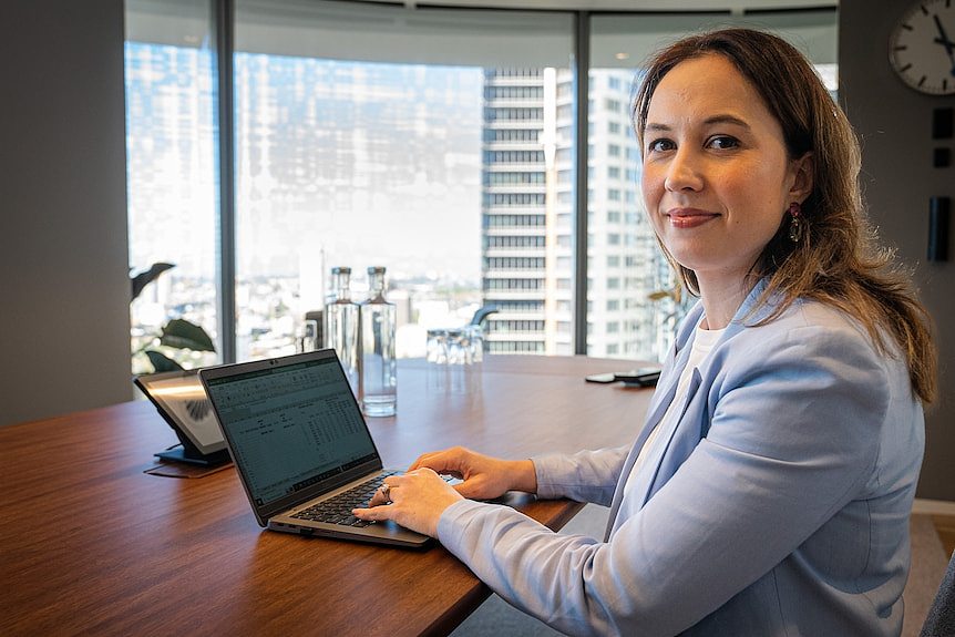 Diana Mousina sits at board table with city views behind her, laptop in front of her