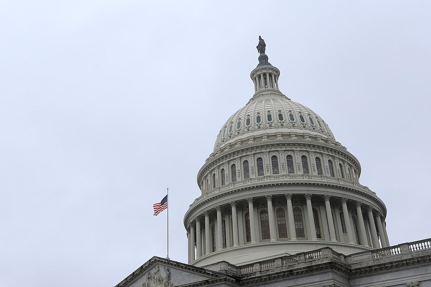 The US Capitol's domed building against a grey sky with a lone US flag flying.
