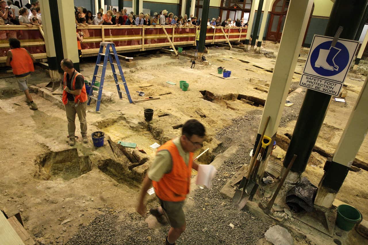身穿反光背心的考古学家在一个小工地上开展工作，工地上挖出几块泥土，人们在路障后面观看。