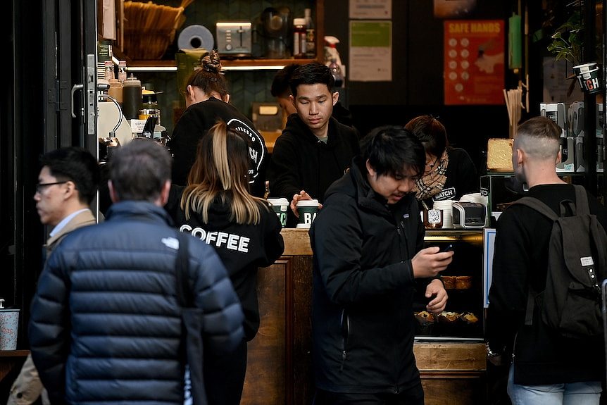 A crowd of customers are a cafe counter, as one person serves coffee, while others look at their phones.