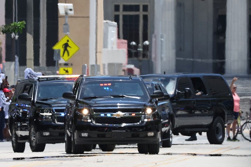 A motorcade of cars gathered on a road with a person walking in the background.